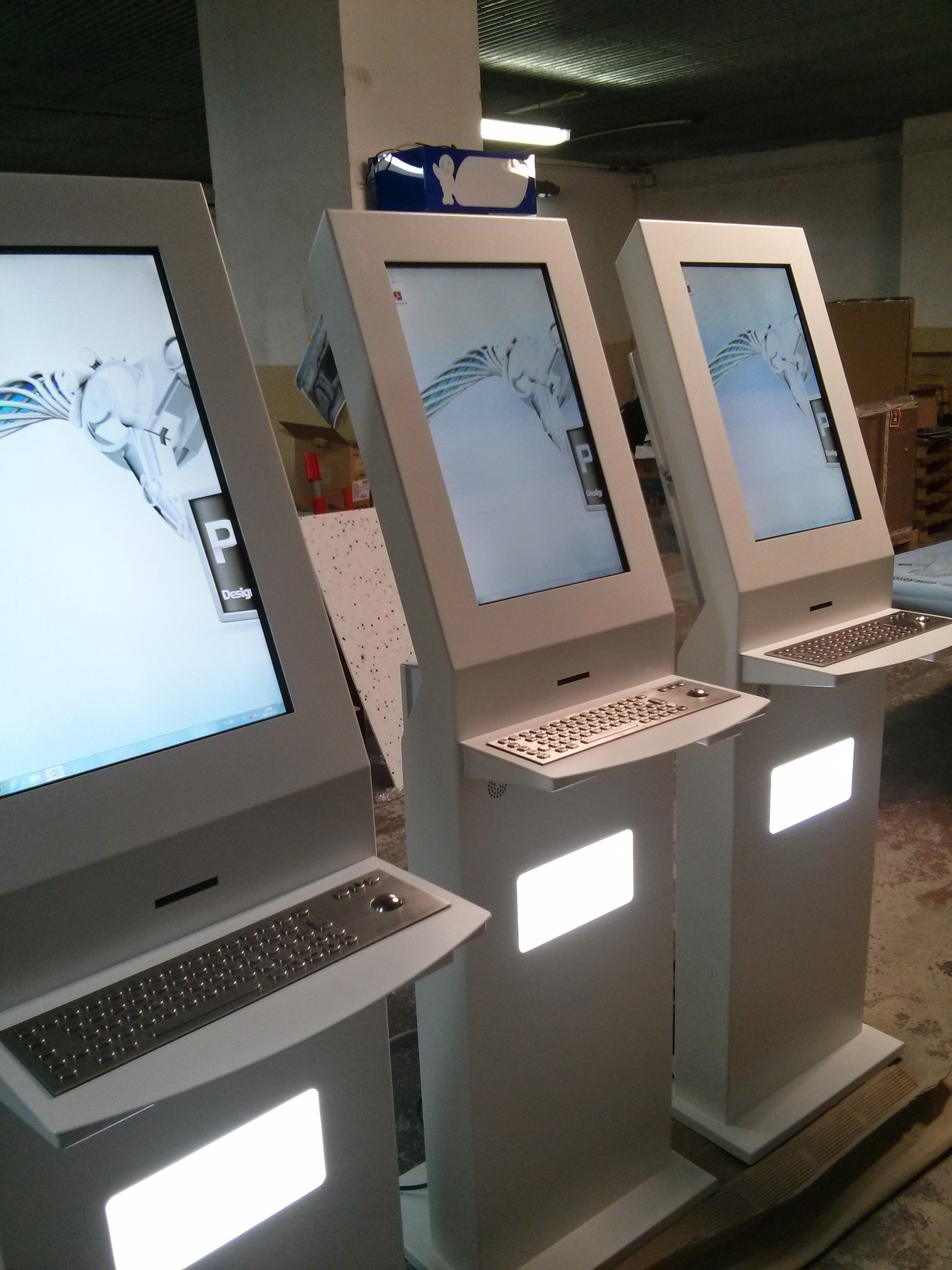 Trzy białe infokioski z pionowymi ekranami dotykowymi i wbudowanymi stalowymi klawiaturami, ustawione obok siebie