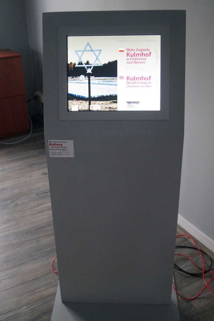 Infokiosk w szarej obudowie prezentujący informacje o obozie zagłady Kulmhof w Chełmnie nad Nerem, ustawiony w pomieszczeniu z drewnianą podłogą.