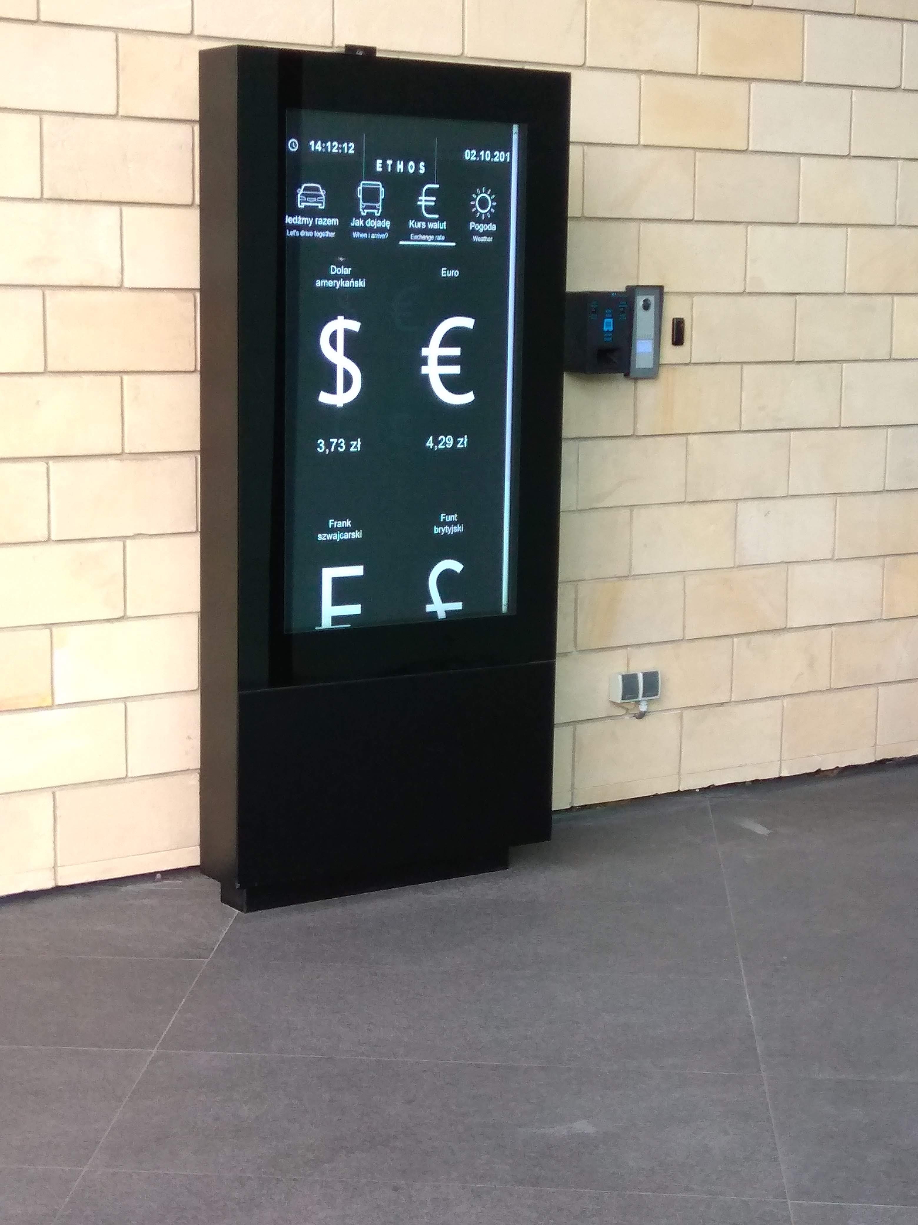 Czarny infokiosk z pionowym ekranem prezentującym informacje o kursach walut, ustawiony przy jasnej kamiennej ścianie w nowoczesnym wnętrzu budynku.