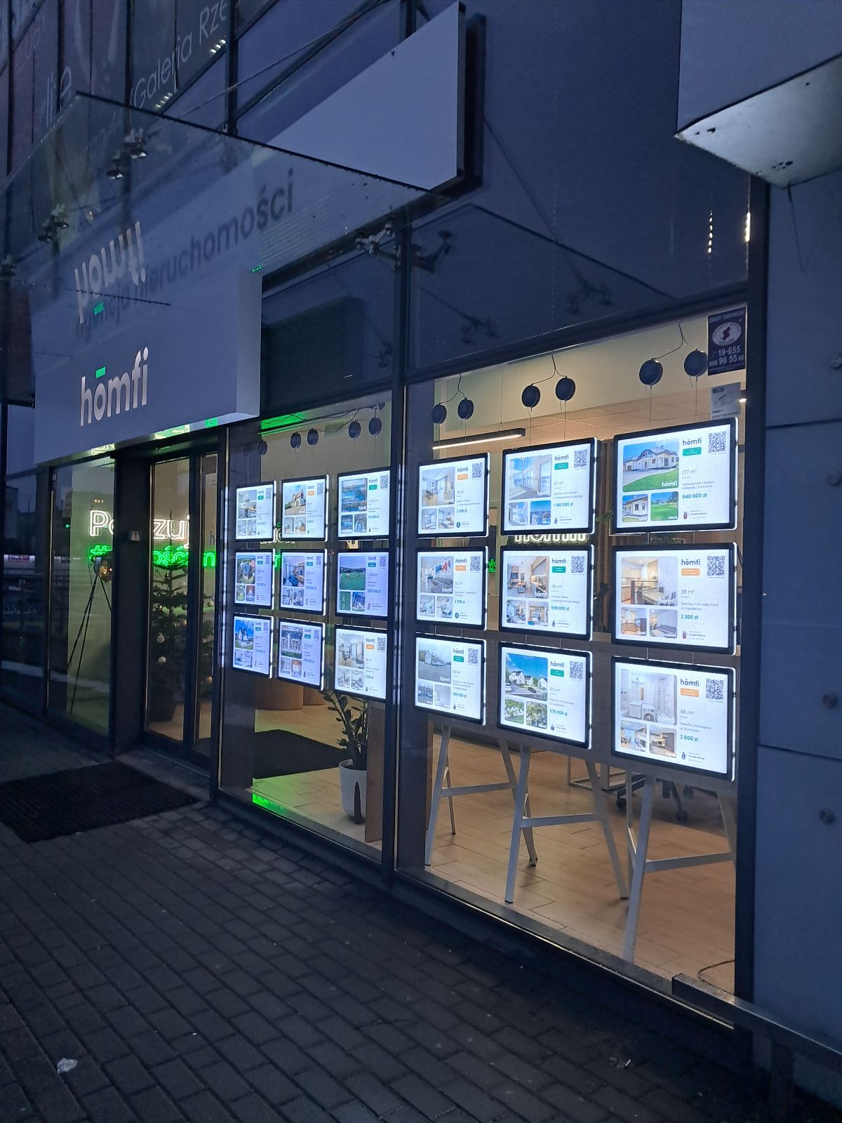 Agencja nieruchomości Homfi z witryną wypełnioną ramkami LED po zmroku