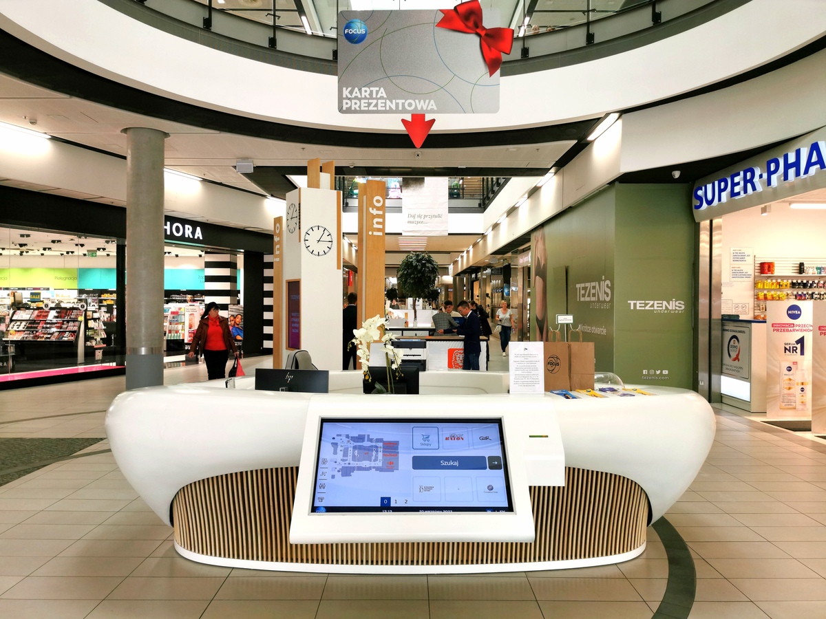 Nowoczesny punkt informacyjny w galerii handlowej, wyposażony w interaktywny ekran dotykowy do nawigacji po obiekcie. Stanowisko obsługi klienta zlokalizowane w centralnej części pasażu.