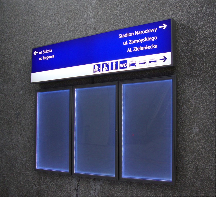 Podświetlana tablica informacyjna na ścianie z oznaczeniami kierunkowymi do kluczowych lokalizacji, takich jak Stadion Narodowy. Poniżej znajdują się trzy gabloty informacyjne z podświetleniem LED.