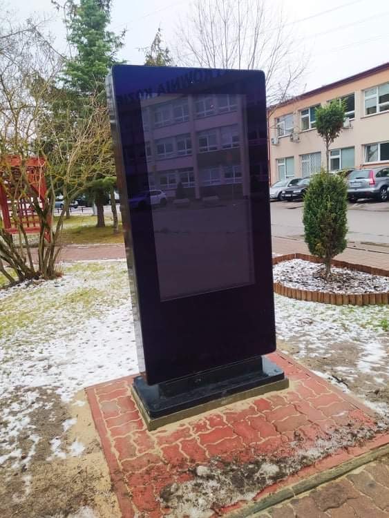 Nowoczesny infokiosk z czarną obudową ustawiony na zewnątrz budynku, na wybrukowanym podeście, w otoczeniu zimowego krajobrazu z lekkim śniegiem.