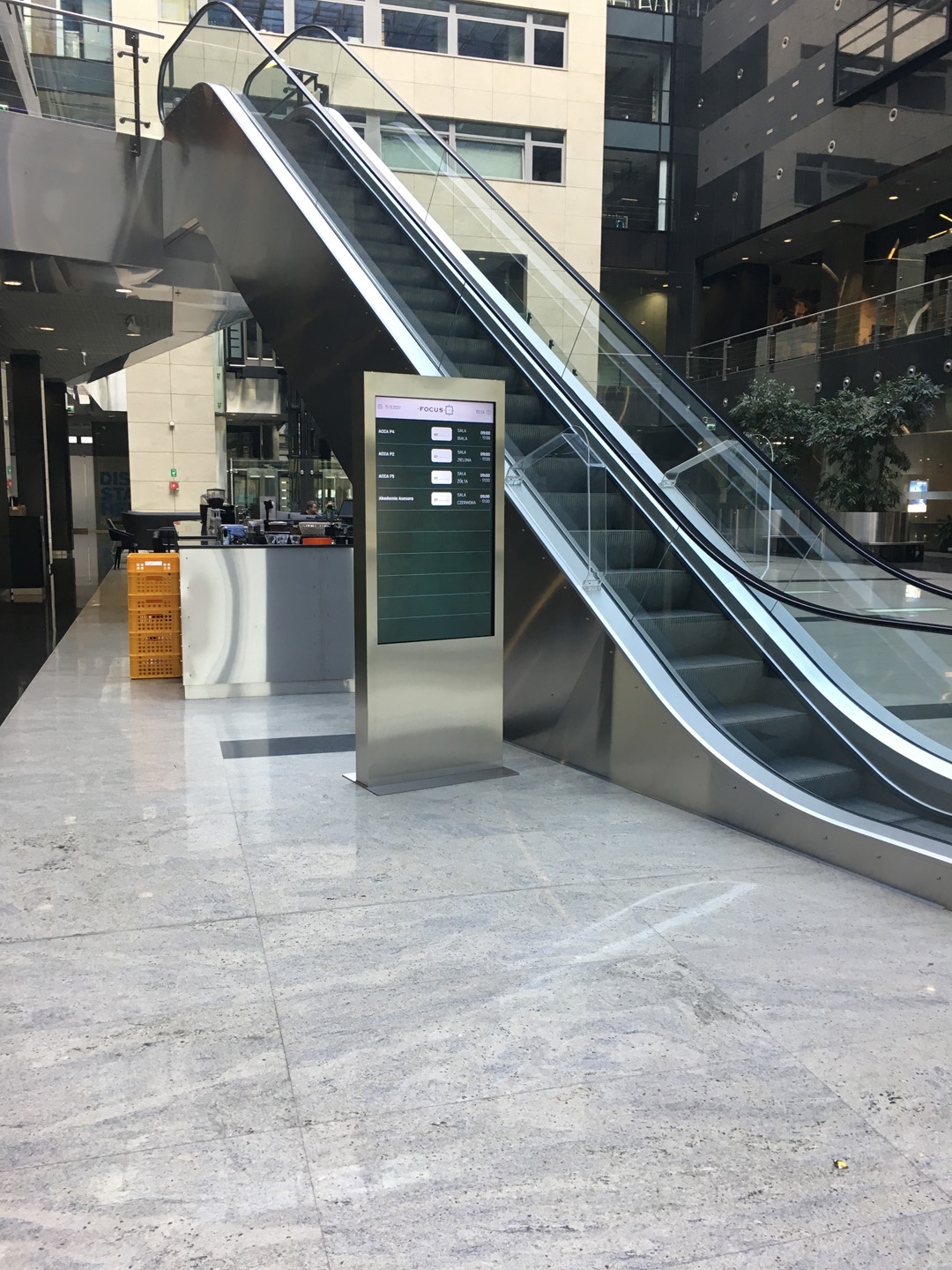 Srebrny infokiosk z ekranem dotykowym wyświetlający informacje o seansach kinowych, ustawiony przy ruchomych schodach w nowoczesnym wnętrzu centrum handlowego.