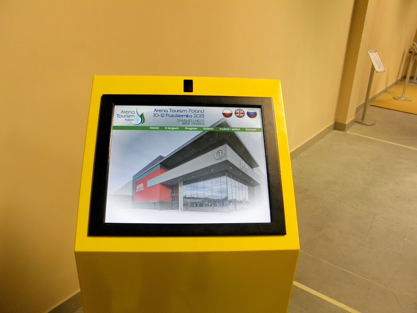 Żółty infokiosk z ekranem dotykowym prezentującym informacje o wydarzeniu Arena Tourism Poland, ustawiony wewnątrz budynku targowego.