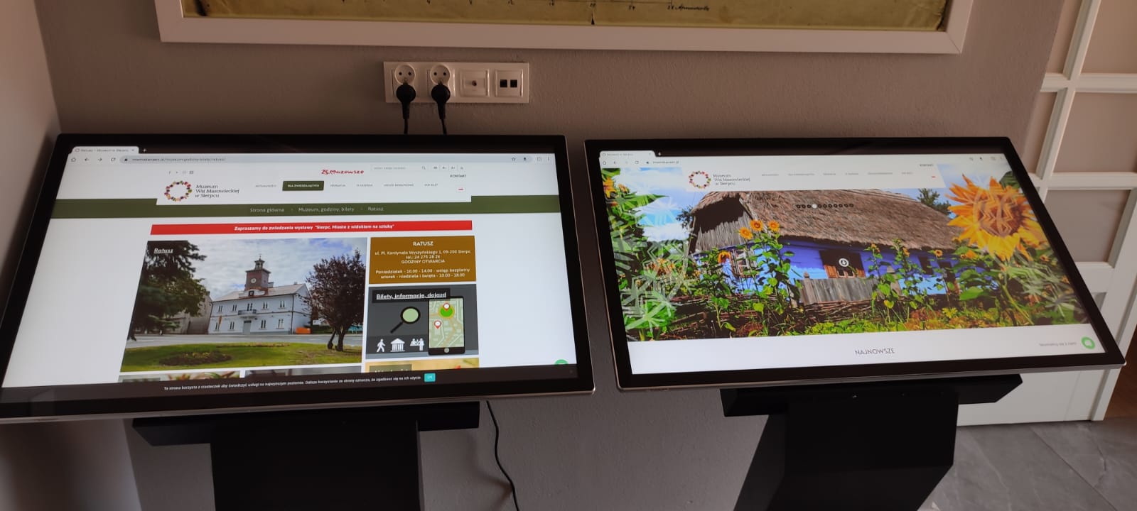 Dwa interaktywne infokioski z dużymi ekranami dotykowymi, prezentujące stronę internetową Muzeum Wsi Mazowieckiej w Sierpcu, ustawione obok siebie przy ścianie wewnętrznej.
