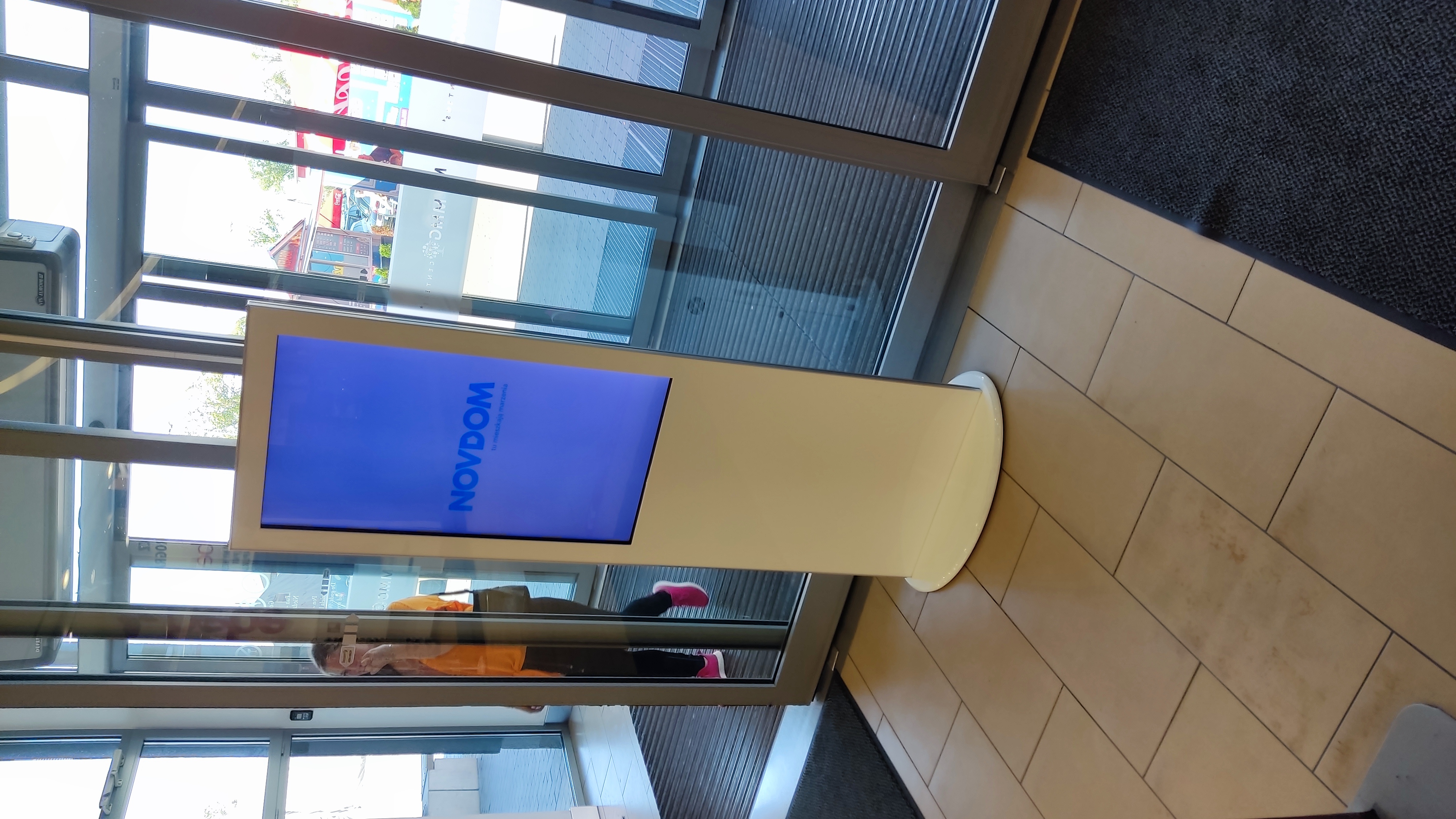 Infokiosk digital signage w galerii handlowej – ekran z ofertą deweloperską przy wejściu do budynku.
