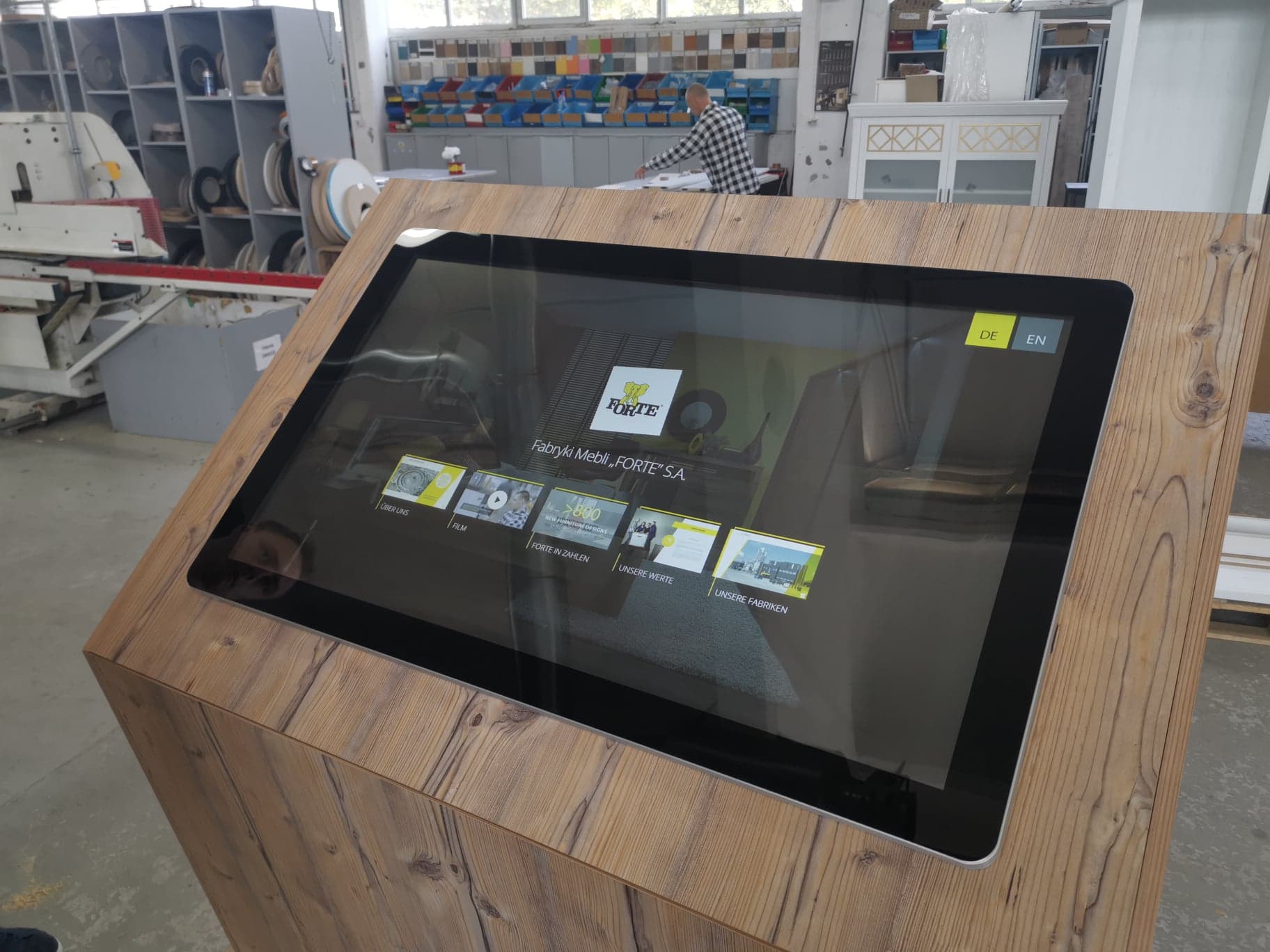 Monitor dotykowy w drewnianej obudowie z interfejsem prezentującym informacje o fabryce mebli FORTE, ustawiony w hali produkcyjnej.