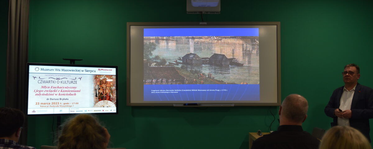 Prelekcja w sali z dwoma ekranami – po lewej monitor z informacją o wydarzeniu kulturalnym Muzeum Wsi Mazowieckiej w Sierpcu, po prawej projektor wyświetlający historyczną ilustrację, obok prowadzący wykład mężczyzna.