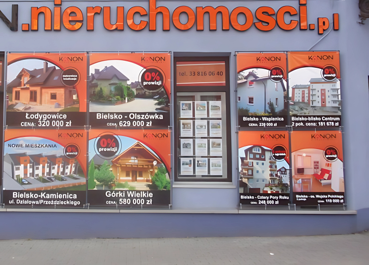 Zdjęcie przedstawia witrynę biura nieruchomości, w której wykorzystano tradycyjne systemy linkowe. W transparentnych kieszeniach akrylowych zawieszonych na stalowych linkach prezentowane są materiały reklamowe dotyczące ofert sprzedaży domów i mieszkań, zawierające zdjęcia, lokalizacje oraz ceny nieruchomości. Całość ekspozycji jest przejrzysta, uporządkowana i estetyczna.