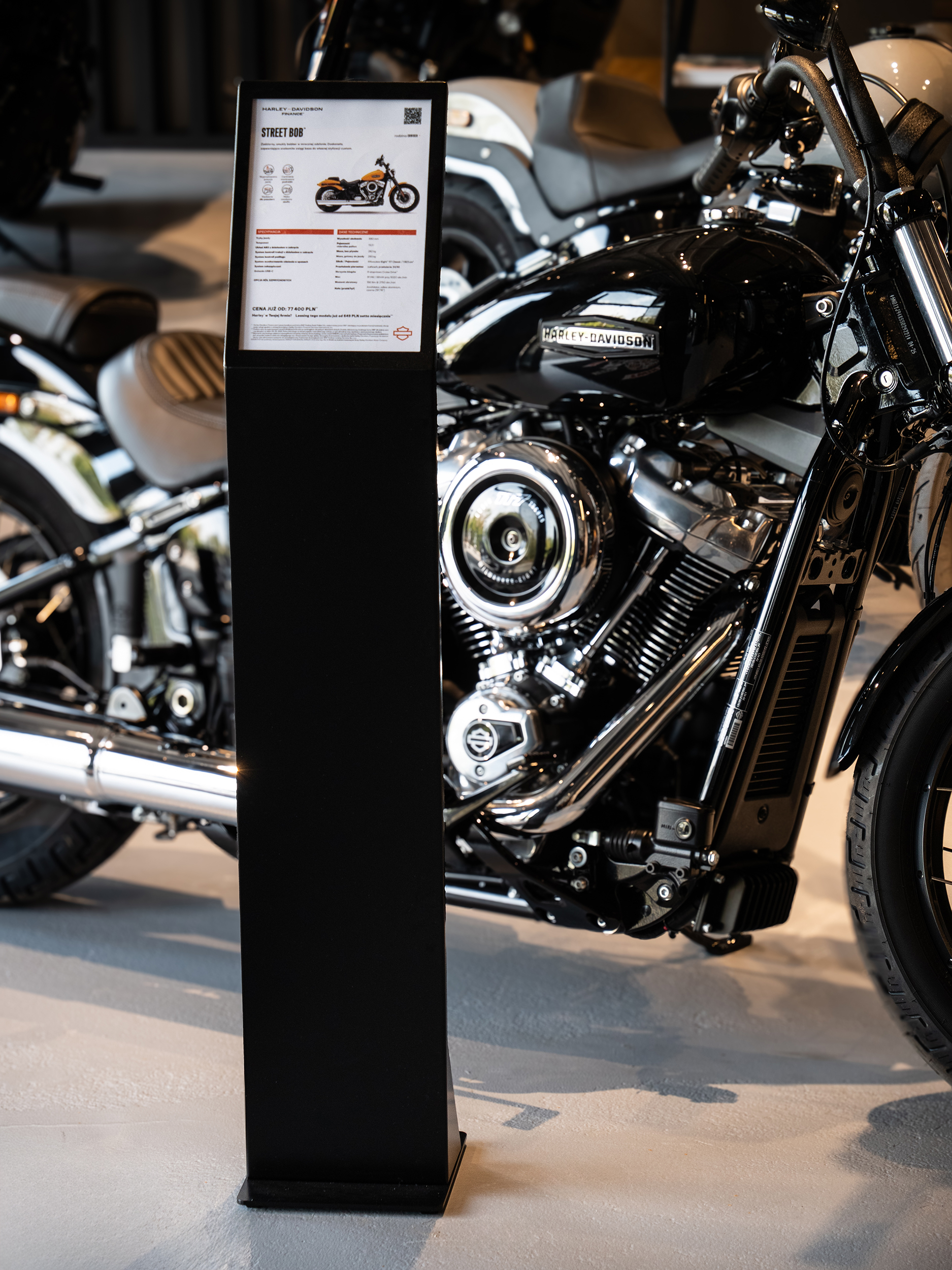 Stojak informacyjny z ulotkami przy motocyklu Harley-Davidson w salonie sprzedaży.