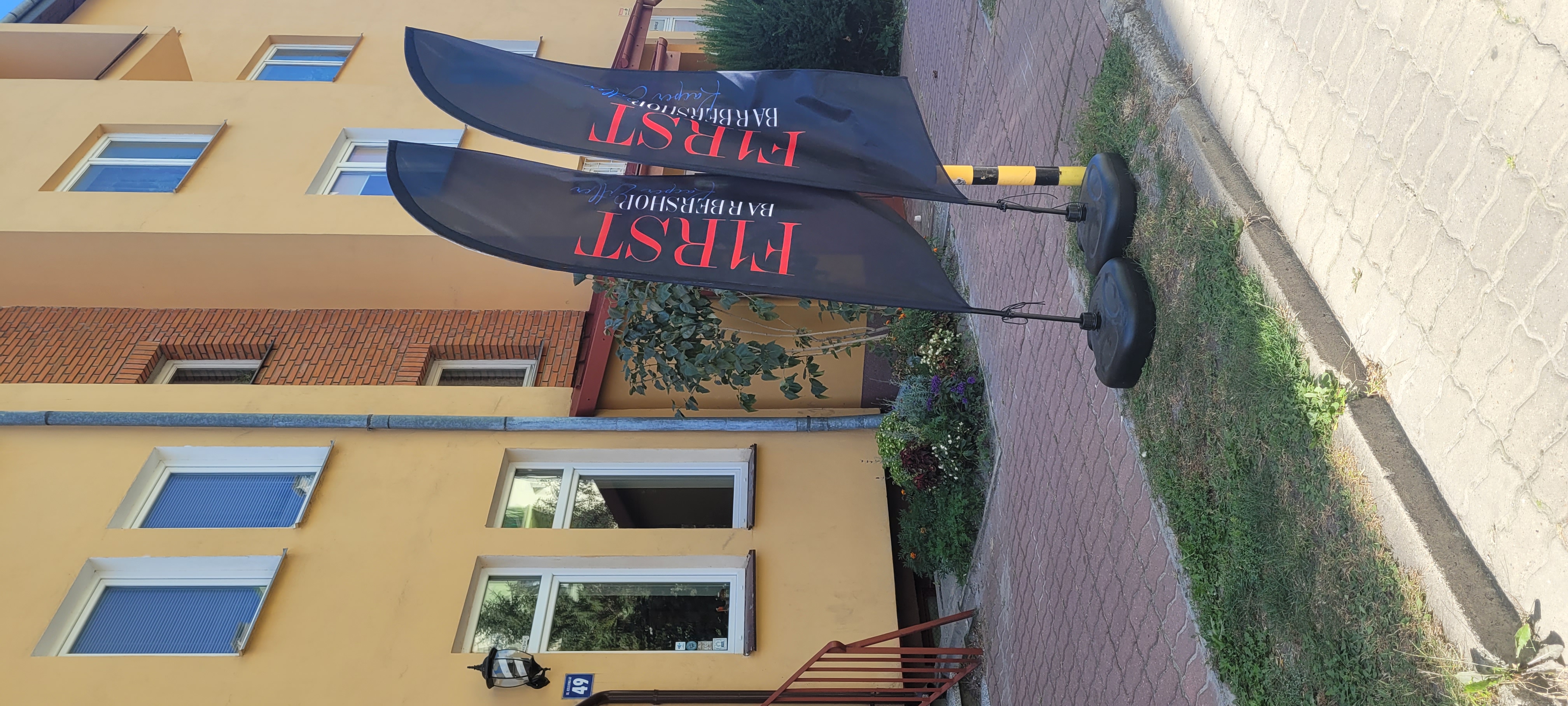 Dwie czarne flagi reklamowe FIRST Barbershop ustawione przy wejściu do salonu.