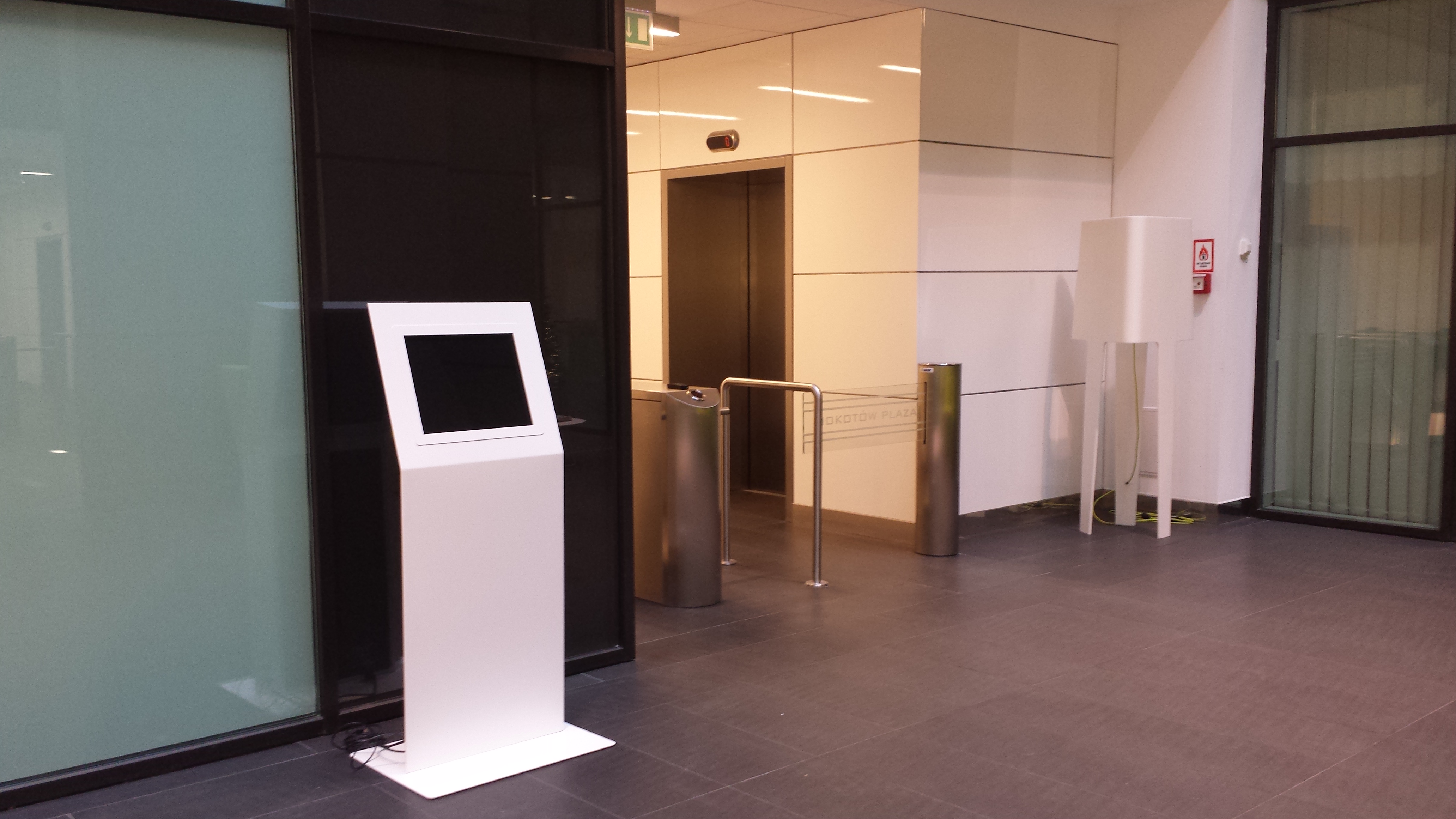 Biały infokiosk z ekranem dotykowym ustawiony w nowoczesnym, eleganckim holu biurowca przy wejściu z kontrolą dostępu.